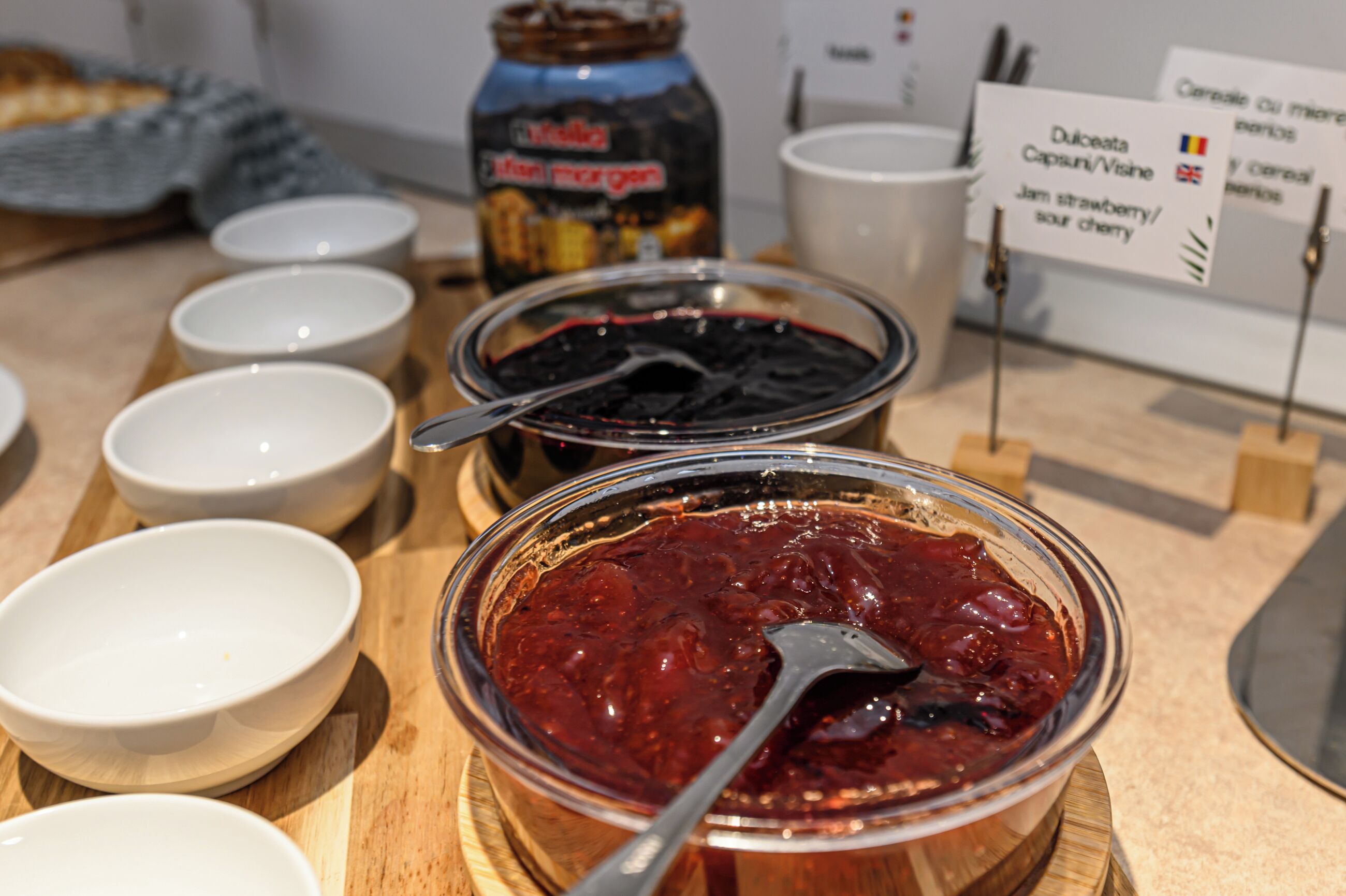 Une table sur laquelle on trouve différents bols contenant divers types de tartinades, un pot de confiture et un panneau indiquant les saveurs, comme la fraise et la cerise.