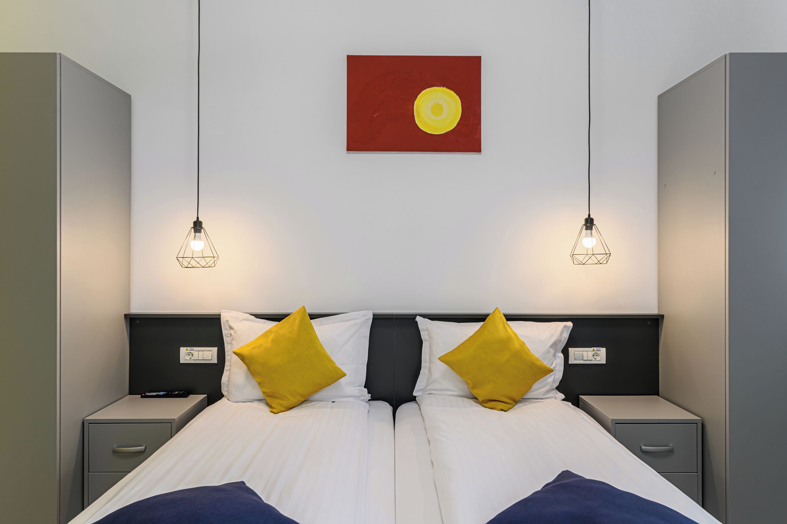 Cama doble con dos mesitas de noche, cada una con una lámpara, una obra de arte roja encima y una pared blanca.