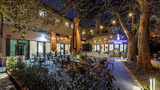 An outdoor patio area with tables and chairs, surrounded by trees and plants, and a building with a sign that reads "JOHANN STRAUSS HOTEL". - Johann Strauss (Bucharest)