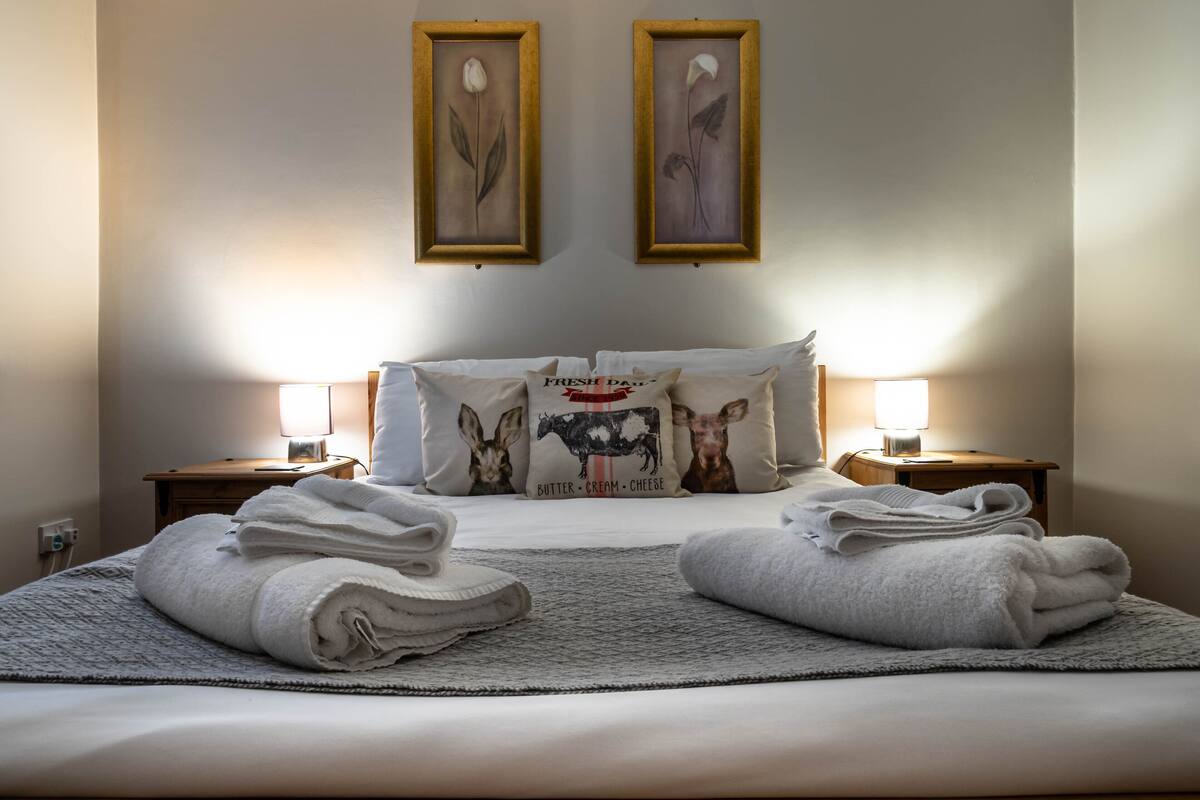Een net opgemaakt bed met twee kussens, een nachtkastje met een lamp, en twee ingelijste foto's aan de muur.
