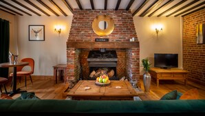 A cozy living room with a brick fireplace, a round mirror, and a television on a wooden table. - The Farmhouse at Fincham (King's Lynn)