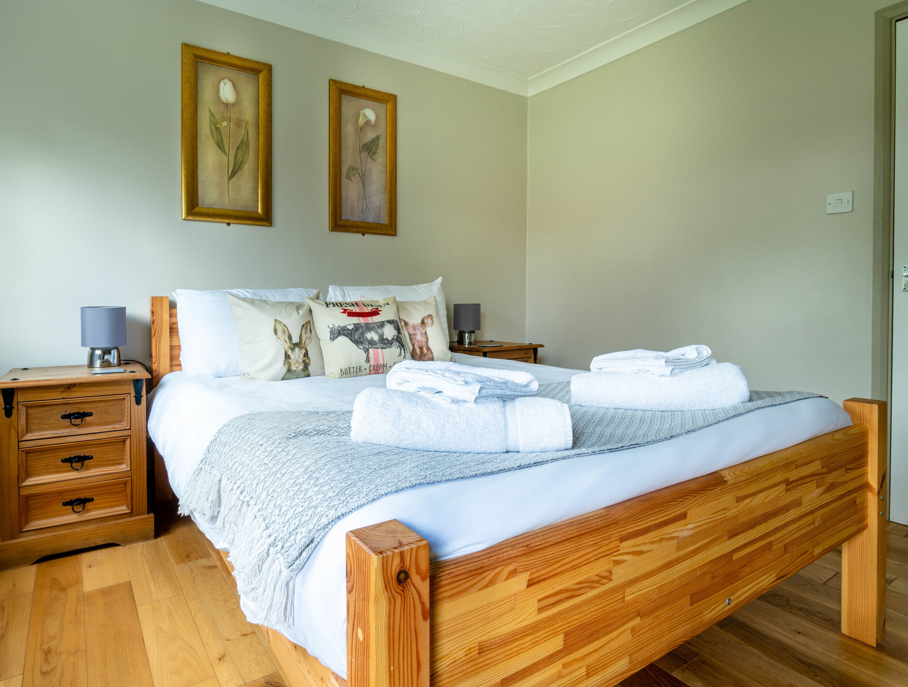 A neatly made bed with white linens and towels, wooden headboard, two bedside tables with lamps, and two framed floral paintings on the wall.