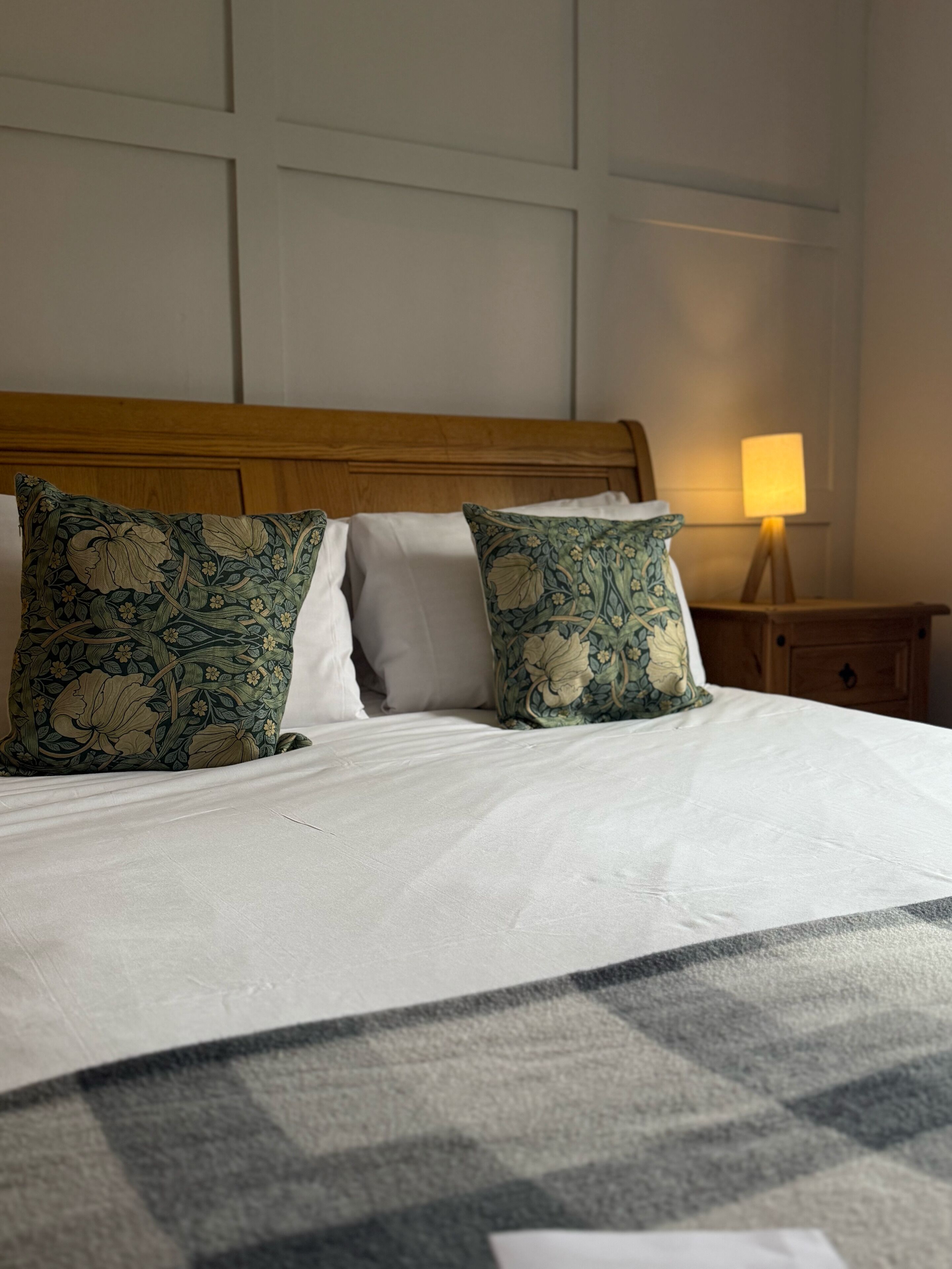 Una cama bien hecha con almohadones estampados de flores, un cabecero de madera y una mesita de noche con una lámpara.