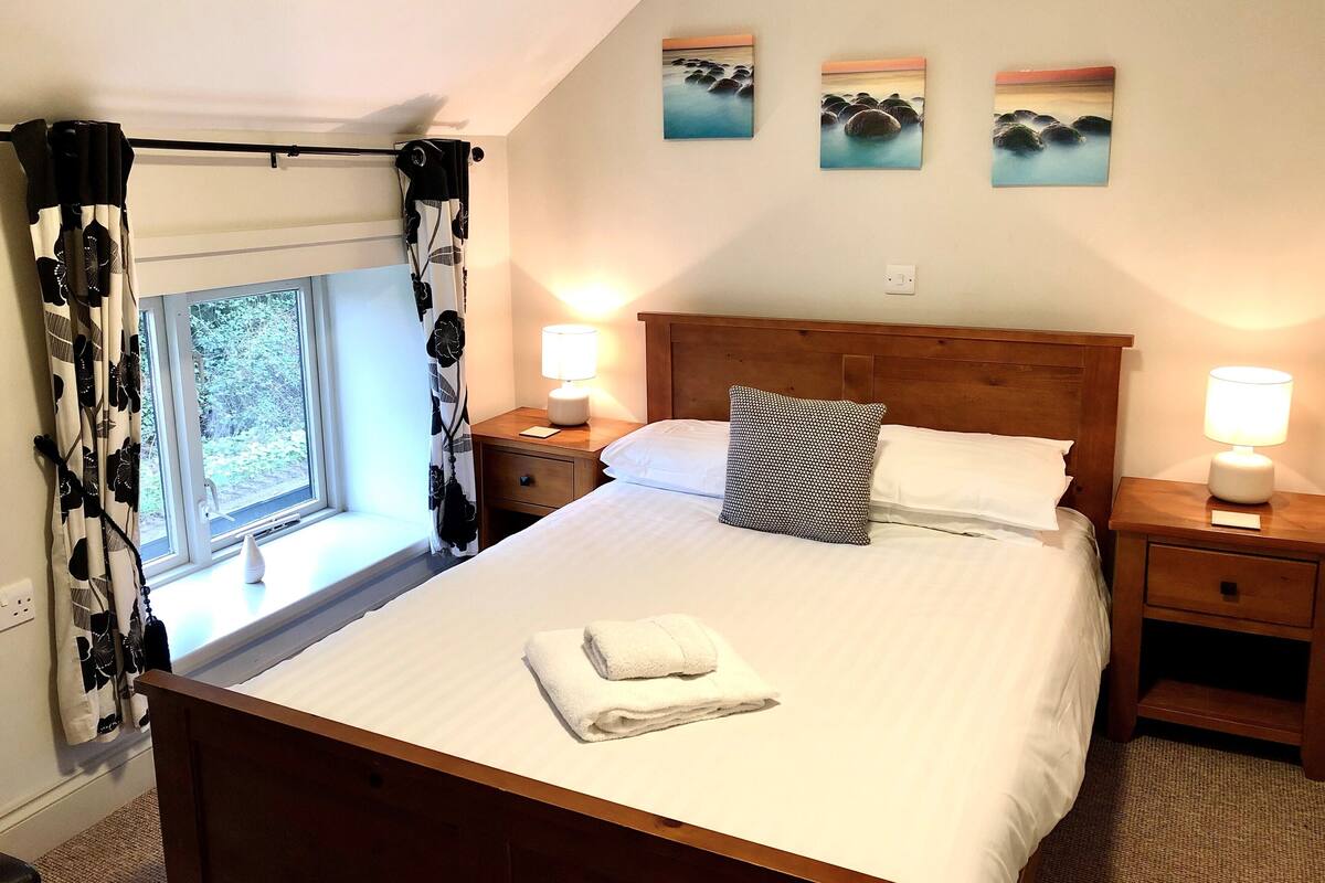 Een net opgemaakt bed met een kussen en een opgevouwen handdoek, een houten hoofdbord, twee nachtkastjes met lampen, een raam met gordijnen, en drie schilderijen aan de muur.