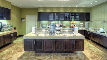 A spacious kitchen with dark wooden cabinets, a large island with various appliances and food items, and a tiled floor.