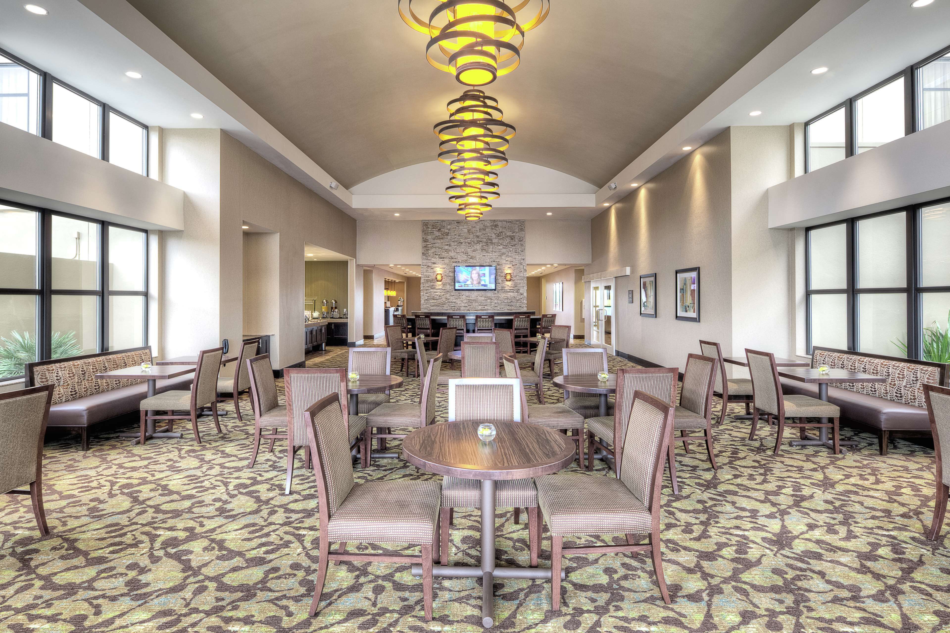 A spacious hotel lobby with a central round table surrounded by chairs, a patterned carpet, and a modern chandelier.