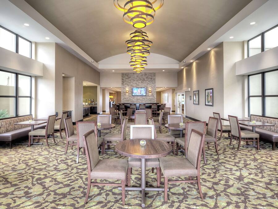 A spacious hotel lobby with a central round table surrounded by chairs, a patterned carpet, and a modern chandelier.