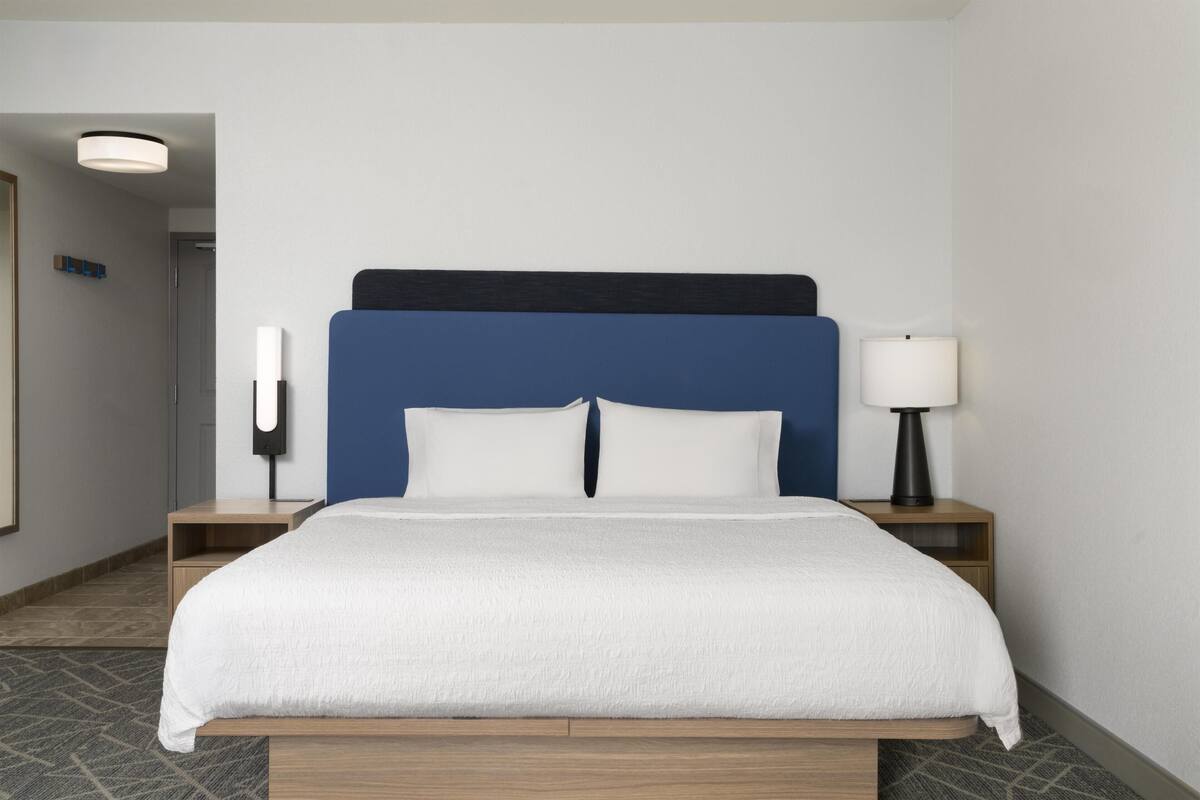 A hotel room with a bed, a nightstand with a lamp, and a wall-mounted light fixture.