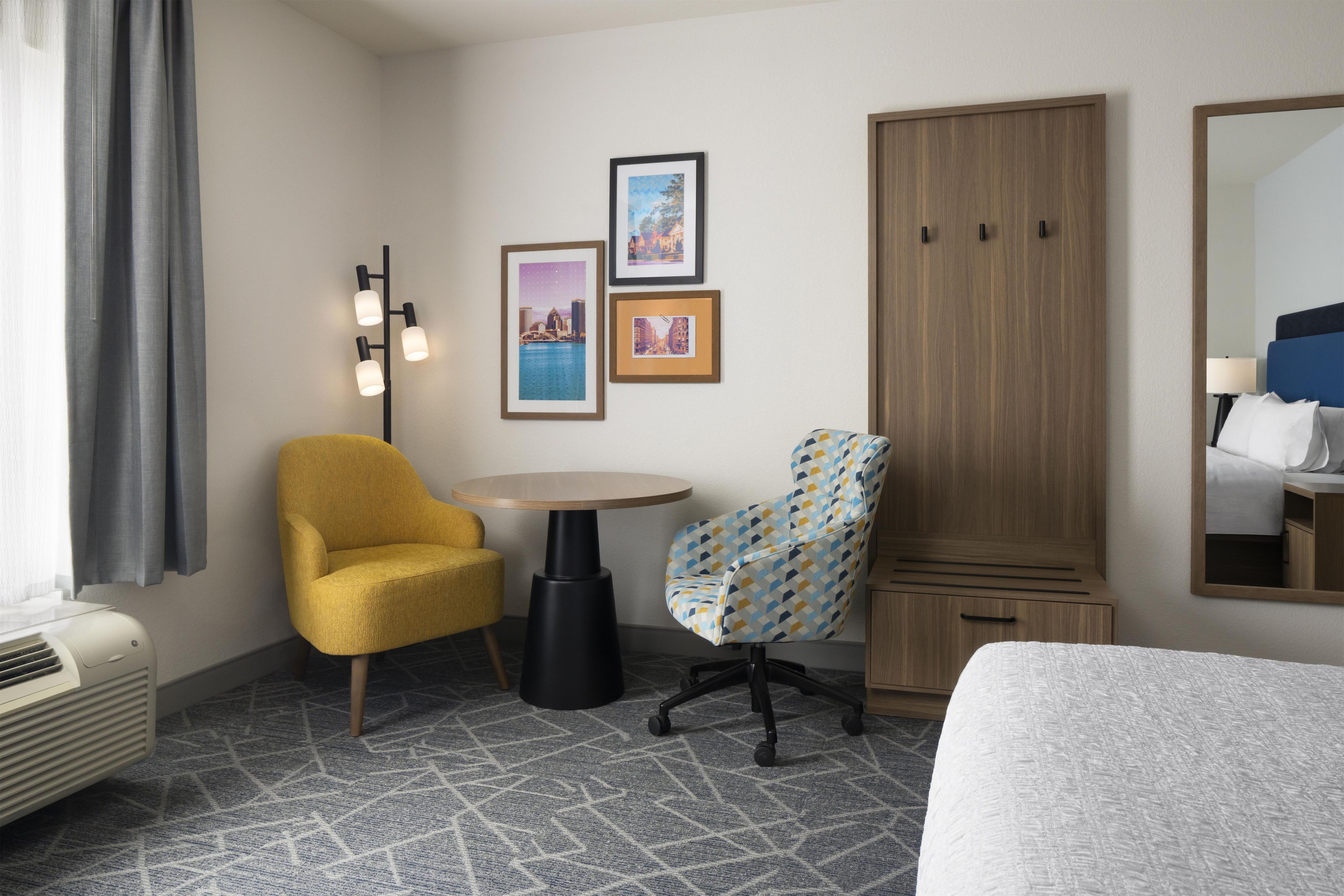 A hotel room with a bed, a yellow armchair, a patterned armchair, a small table, a wardrobe, and a framed picture on the wall.