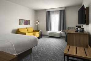 Quarto de hotel com cama, sofá, escrivaninha, televisão, janela com cortinas e radiador.