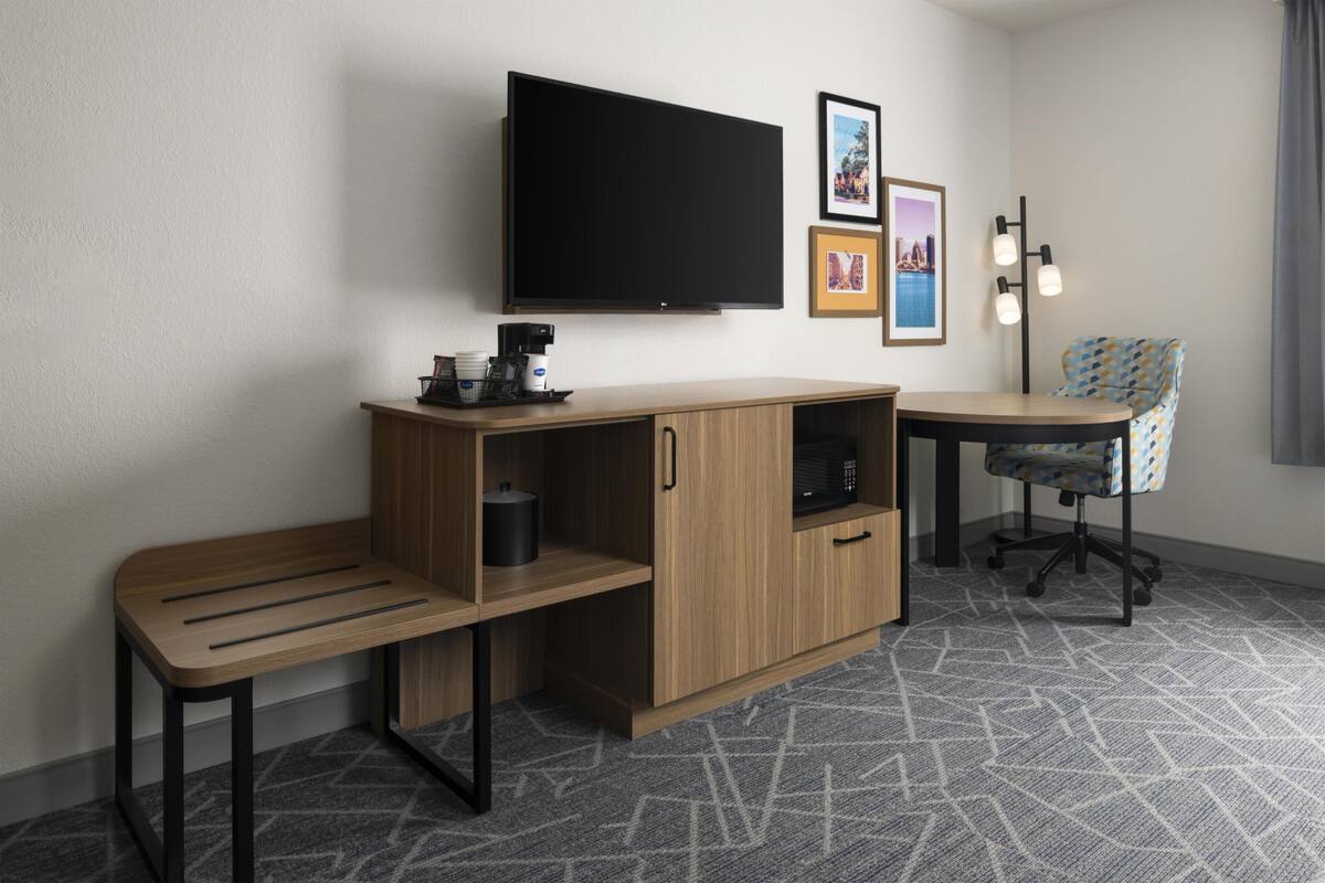 A hotel room with a flat-screen TV mounted on the wall, a wooden desk with a chair, a small cabinet, and a patterned carpet.