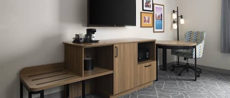 A compact hotel room with a flat-screen TV, a small kitchenette, a desk, a chair, and a patterned carpet.