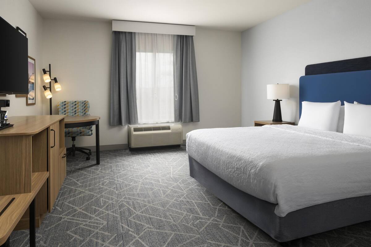 A hotel room with a bed, desk, chair, air conditioning unit, window with curtains, and a television.