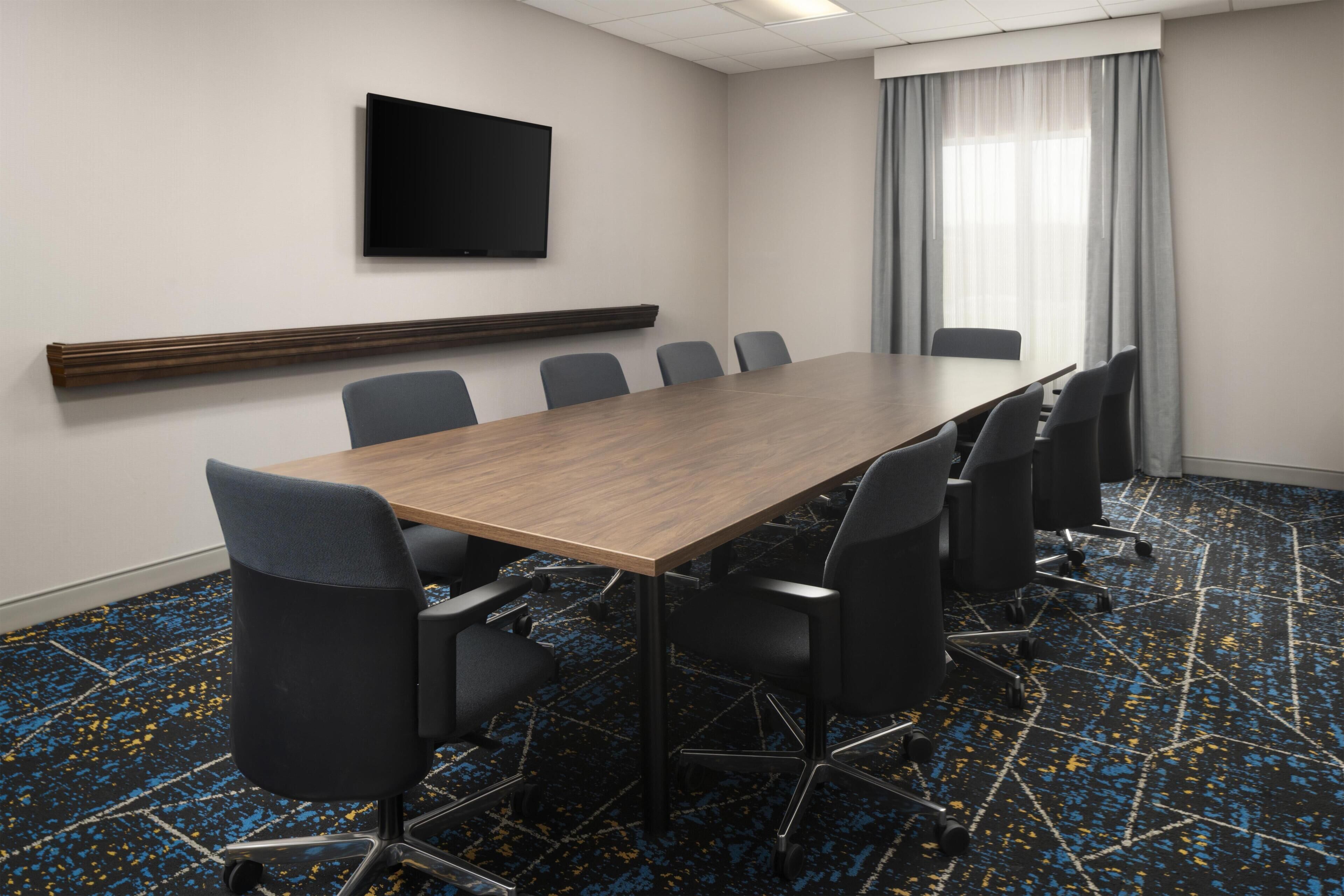 Uma sala de conferências com uma mesa comprida, seis cadeiras, uma TV de tela plana e uma janela com cortinas.
