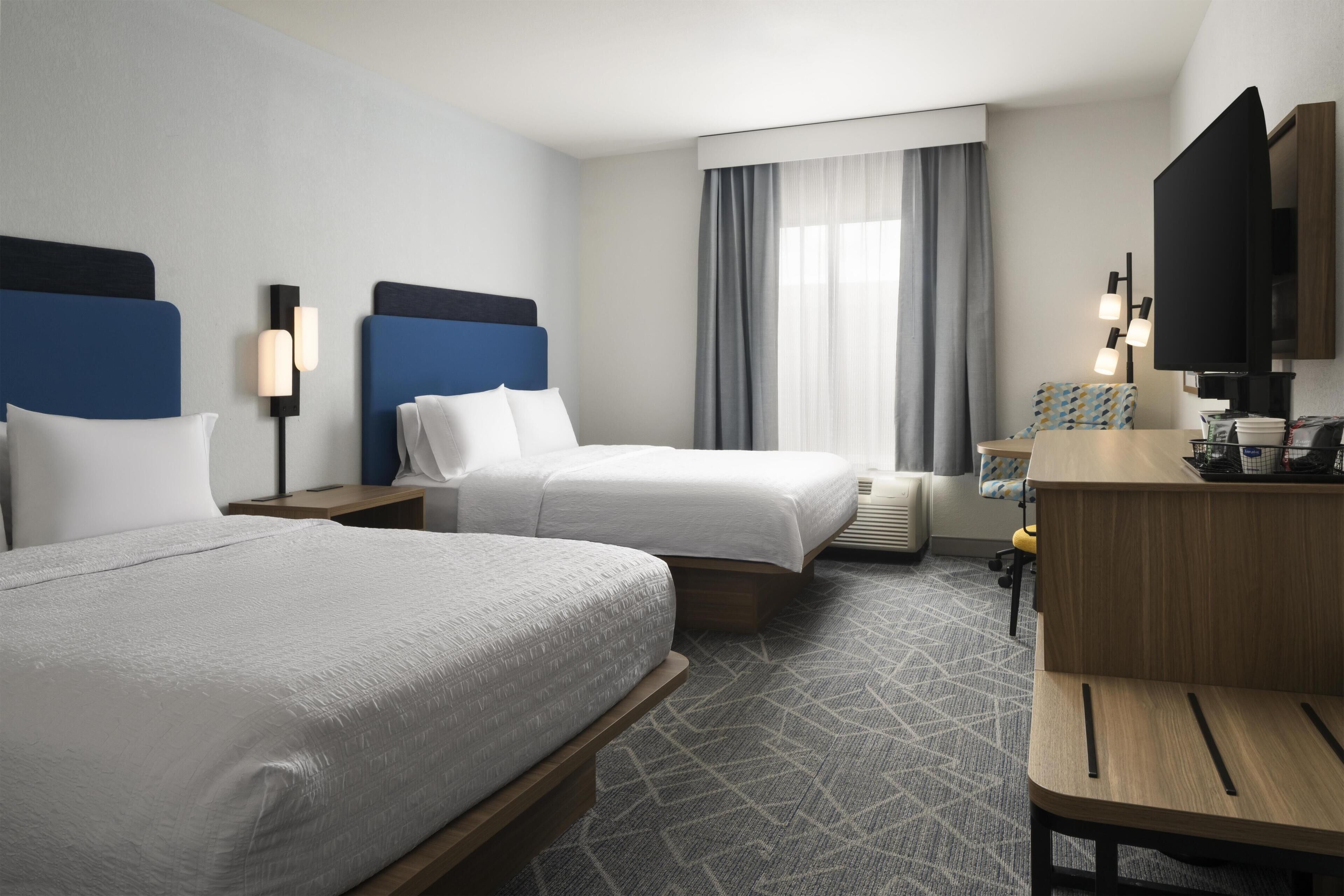 A hotel room with two beds, a desk, a television, and a window with curtains.