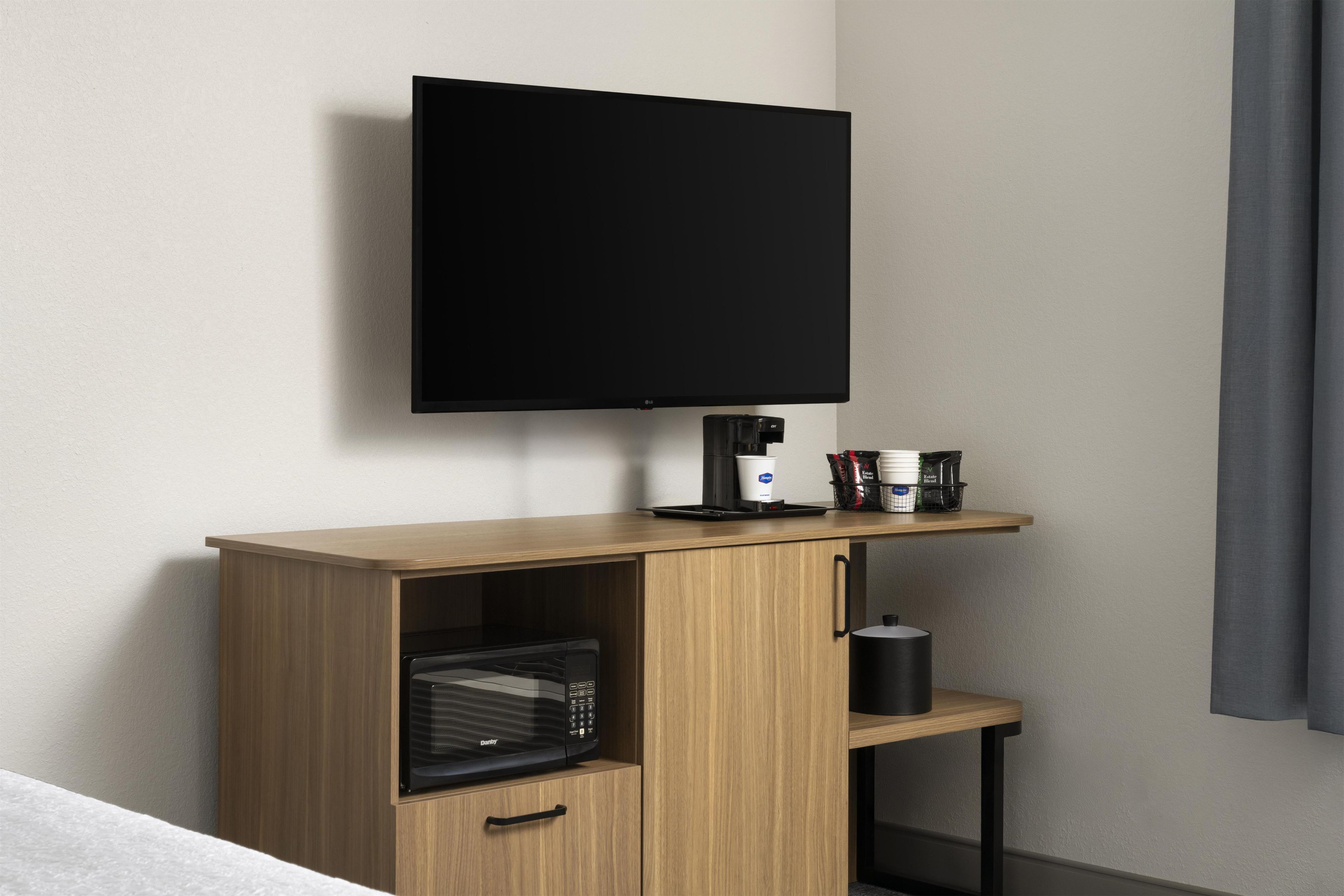 Habitación de hotel con televisor de pantalla plana montado en la pared, microondas en un mueble de madera, cafetera y una mesita con una taza y una bolsa de aperitivos.