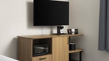 Habitación de hotel con televisor de pantalla plana montado en la pared, microondas en un mueble de madera, cafetera y una mesita con una taza y una bolsa de aperitivos.
