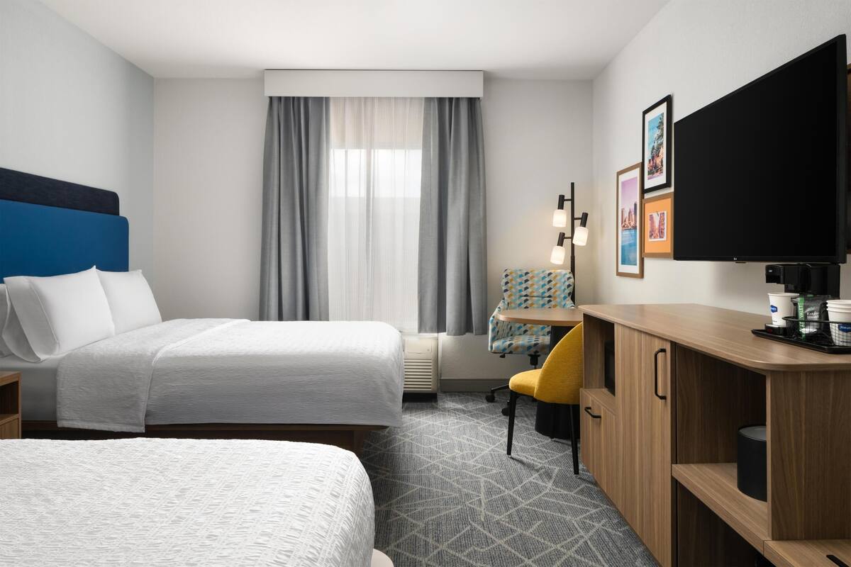 A hotel room with two beds, a desk, a chair, a flat-screen TV, and a window with curtains.