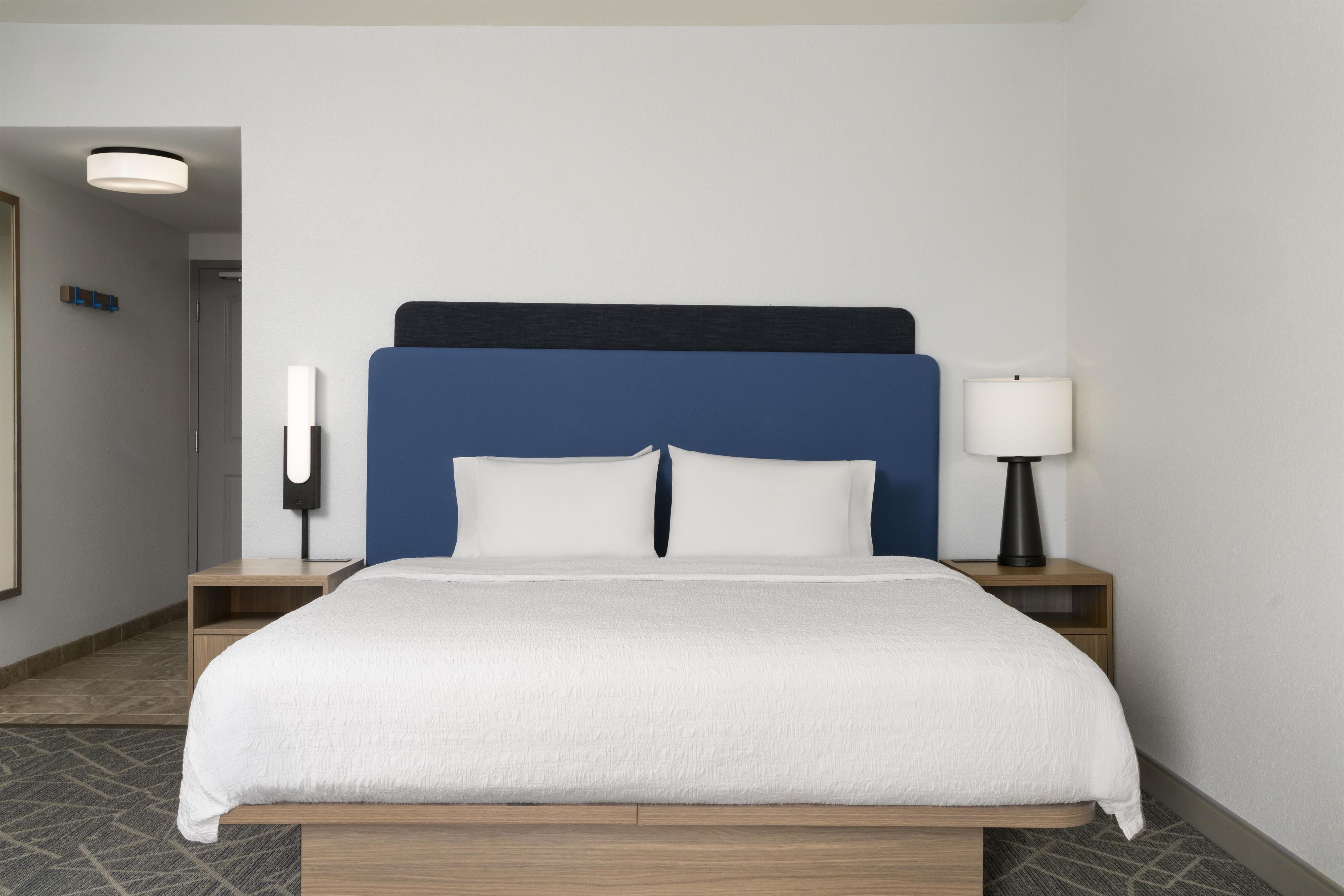 A hotel room with a bed, a nightstand, a lamp, and a wall-mounted light fixture.