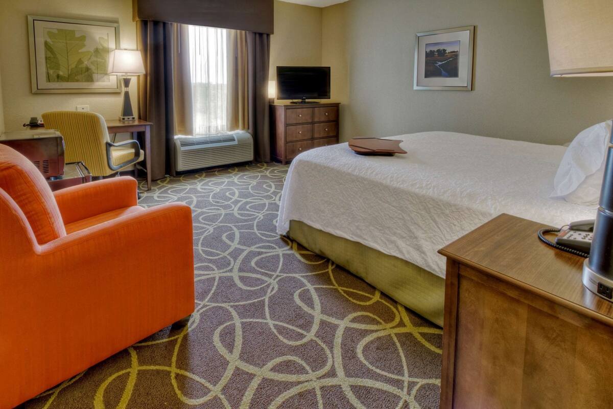A hotel room with a bed, an orange chair, a desk with a telephone, a TV, and a window with curtains.