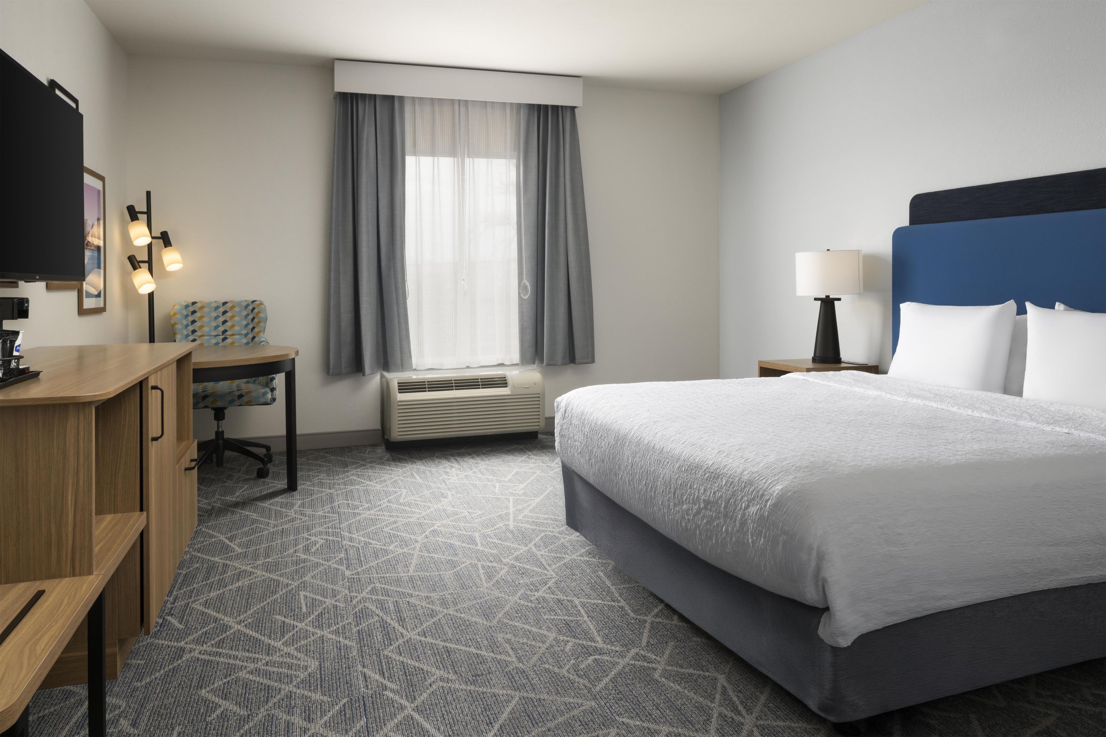 Habitación de hotel con cama, escritorio, silla, unidad de aire acondicionado, ventana con cortinas y televisor.