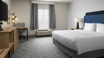 Habitación de hotel con cama, escritorio, silla, unidad de aire acondicionado, ventana con cortinas y televisor.