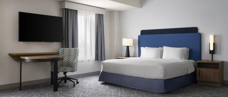 Quarto de hotel com cama, escrivaninha, cadeira, televisão e janela com cortinas.