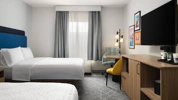 A hotel room with two beds, a desk with a coffee machine, a TV, and a window with curtains.