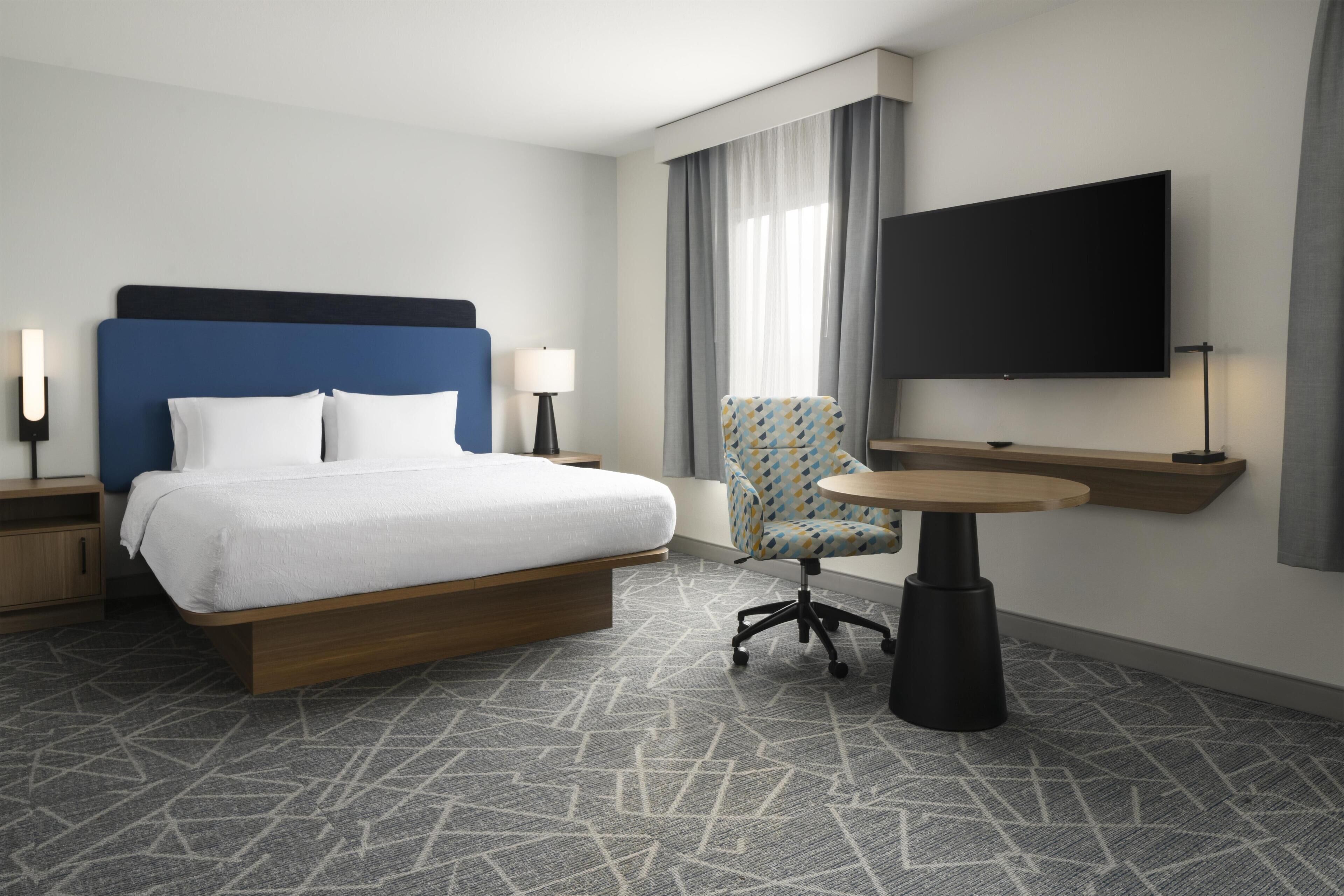 A hotel room with a bed, a chair, a table, a TV, and a lamp.