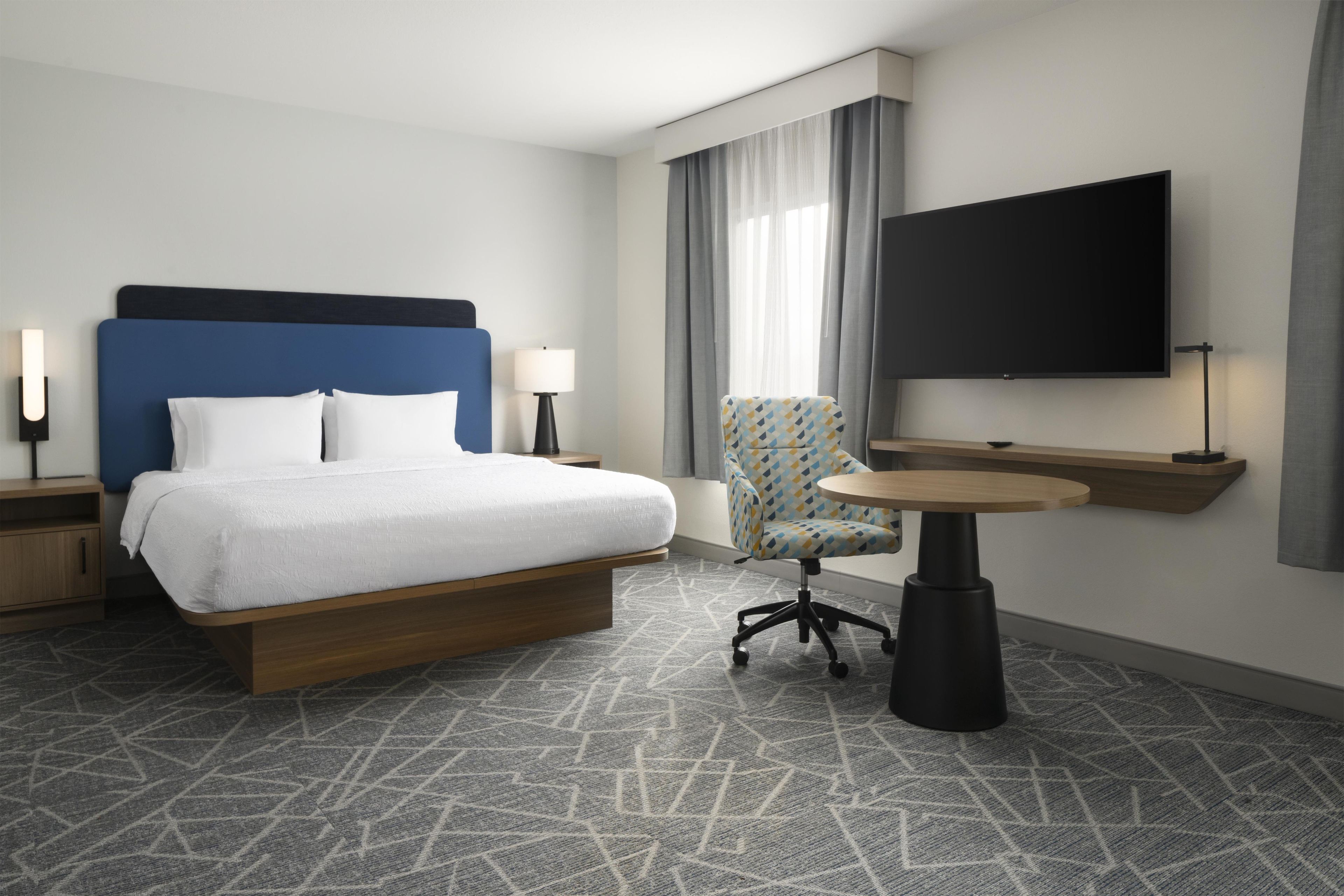 Une chambre d’hôtel avec un lit, une chaise, une petite table, un téléviseur et une table de chevet.