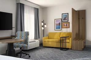 A hotel room with a flat-screen TV, a patterned armchair, a yellow sofa, a small table, and a wooden cabinet.