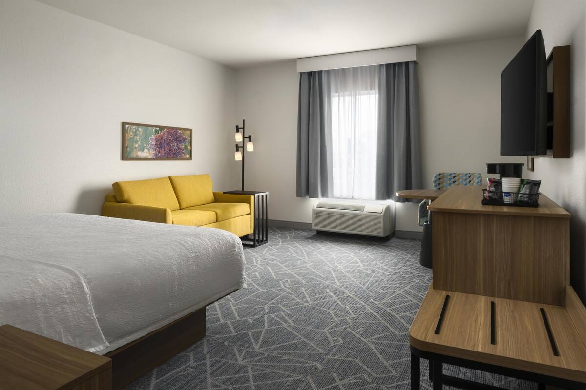 A hotel room with a bed, a yellow armchair, a desk, a flat-screen TV, and a large window with curtains.