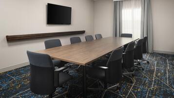 A conference room with a long table, six chairs, a flat-screen TV on the wall, and a window with curtains.