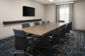 A conference room with a long table, six chairs, a flat-screen TV on the wall, and a window with curtains.