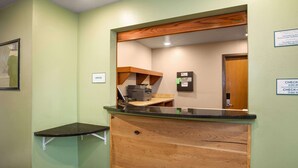 Un espace d’accueil avec un comptoir en bois, un petit bureau et un miroir. Des panneaux indiquent les heures d’ouverture et les heures d’arrivée et de départ.