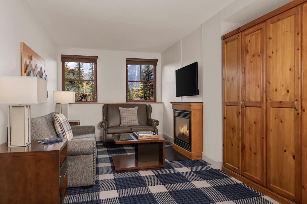 A living room with a sofa, coffee table, and fireplace. There are windows with a view of trees and a television mounted on the wall.