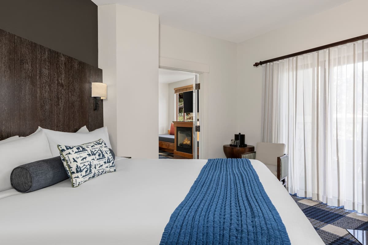 A hotel room with a large bed, a wooden headboard, a bedside table with a lamp, and a view of a fireplace through an open door.