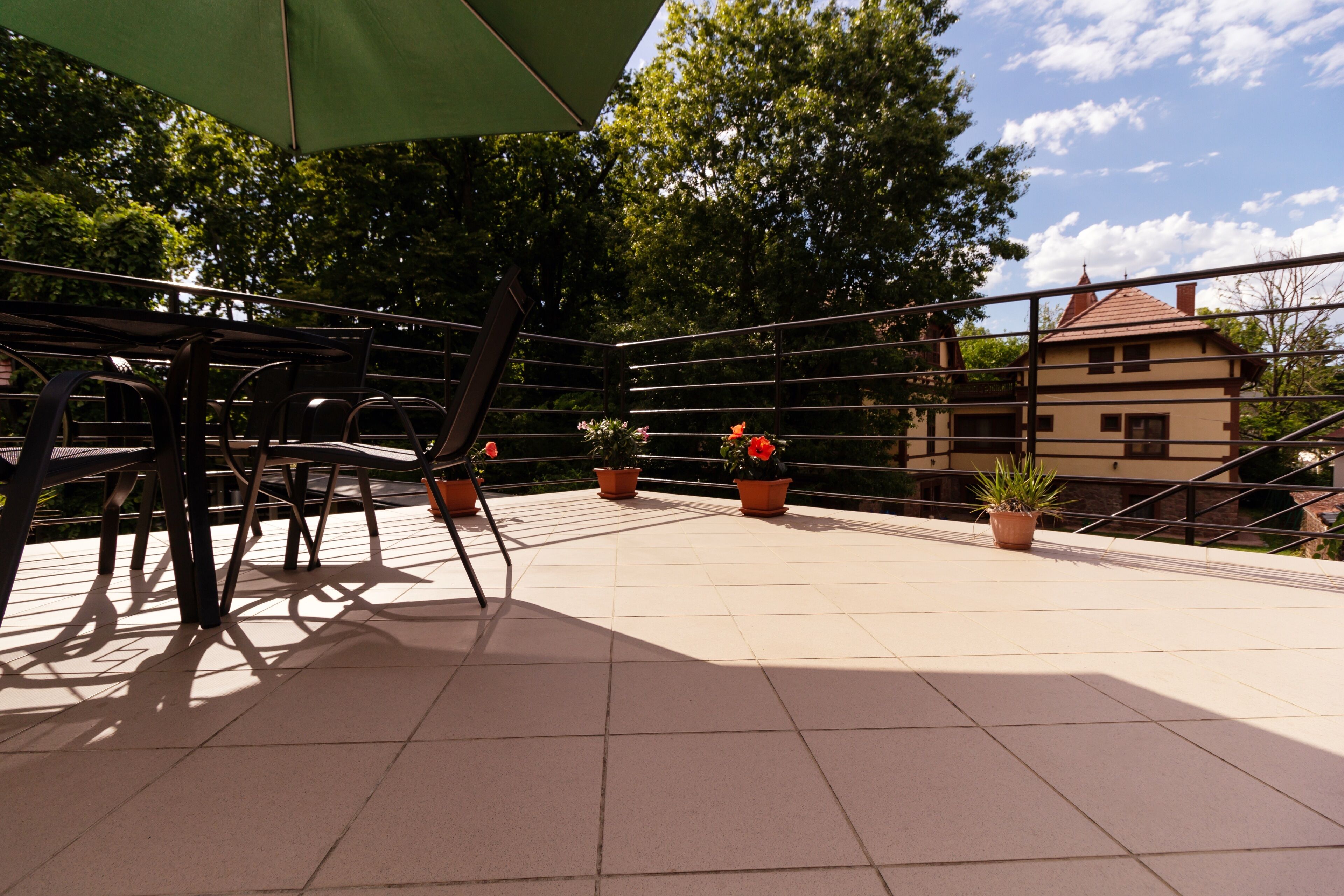 A terrace with chairs and potted plants, a green umbrella, and a view of trees and buildings in the background.