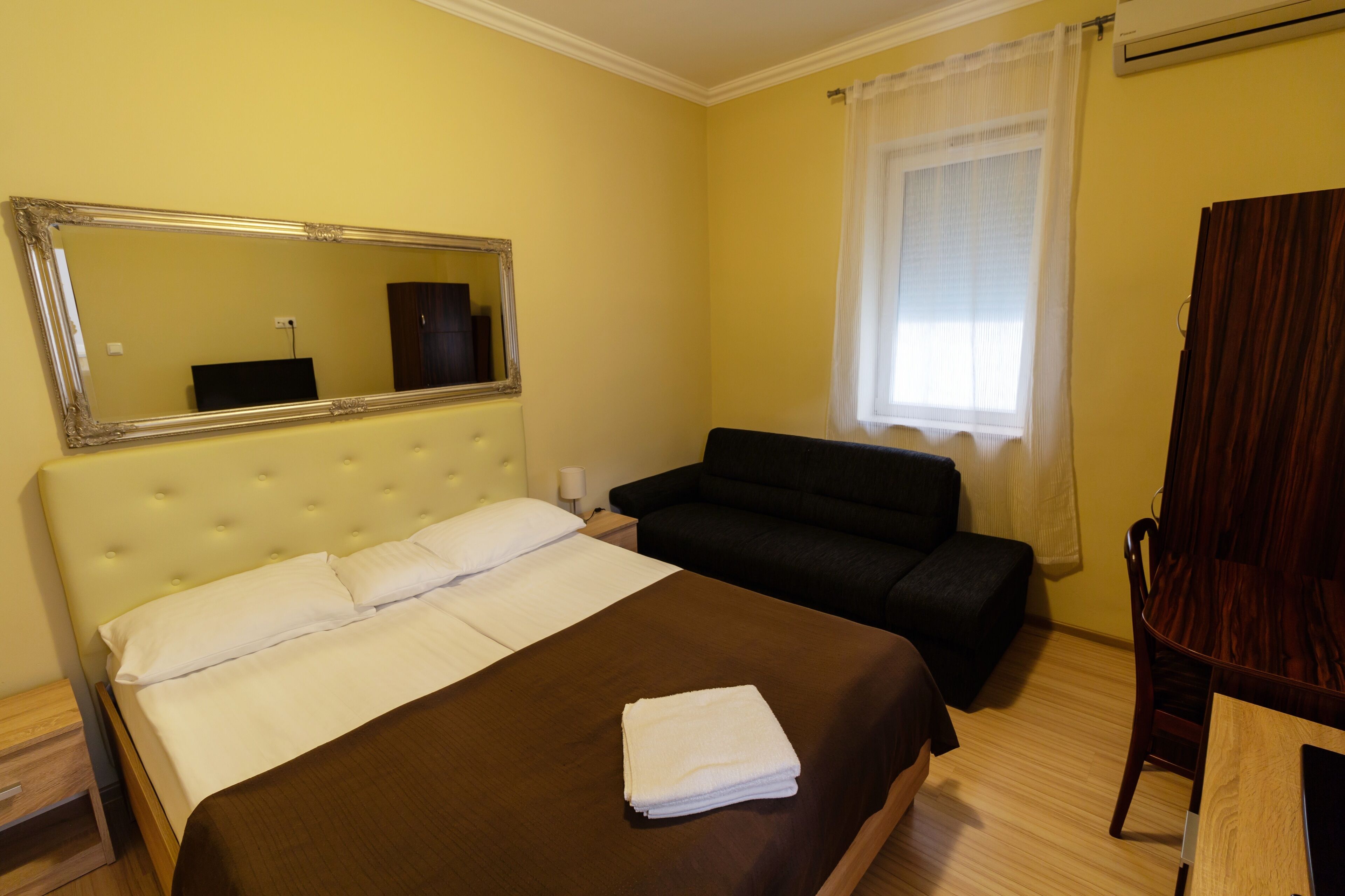 Camera d'albergo con un letto grande, un divano nero, una sedia di legno, uno specchio e una finestra con tende.