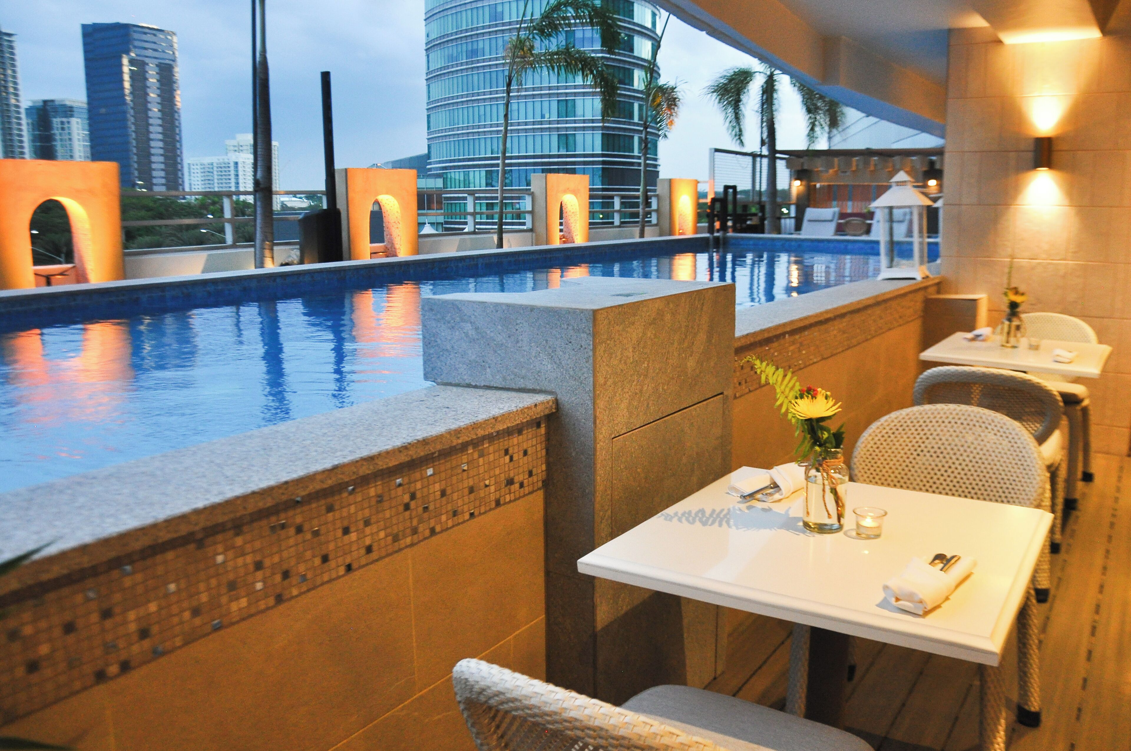 A rooftop pool area with a view of city buildings, a dining table with chairs, and a small white gazebo.