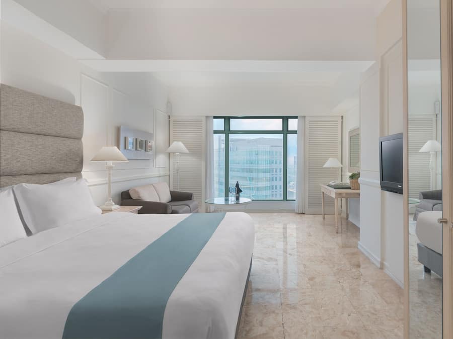 Quarto de hotel moderno com uma cama grande, uma mesa de trabalho, uma televisão e vista para o horizonte urbano pela janela.