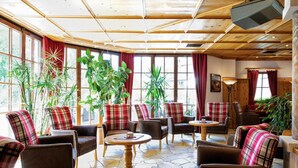 A cozy lounge area with plaid-patterned chairs and tables, large windows with red curtains, and various indoor plants. - Hotel Appartement Similaun (Soelden)
