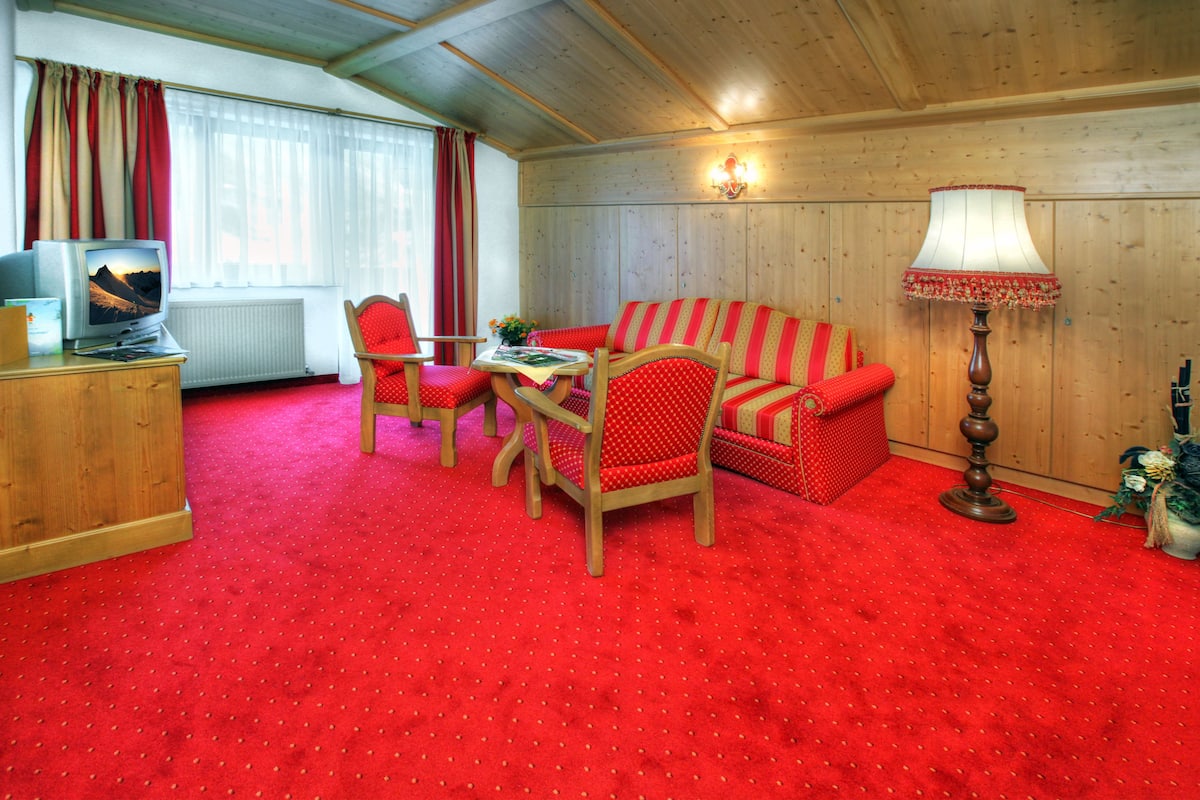 Ein Zimmer mit rotem Teppich, Holzwänden, einer Couch, zwei Stühlen, einem Tisch, einer Lampe und einem Fernseher.
