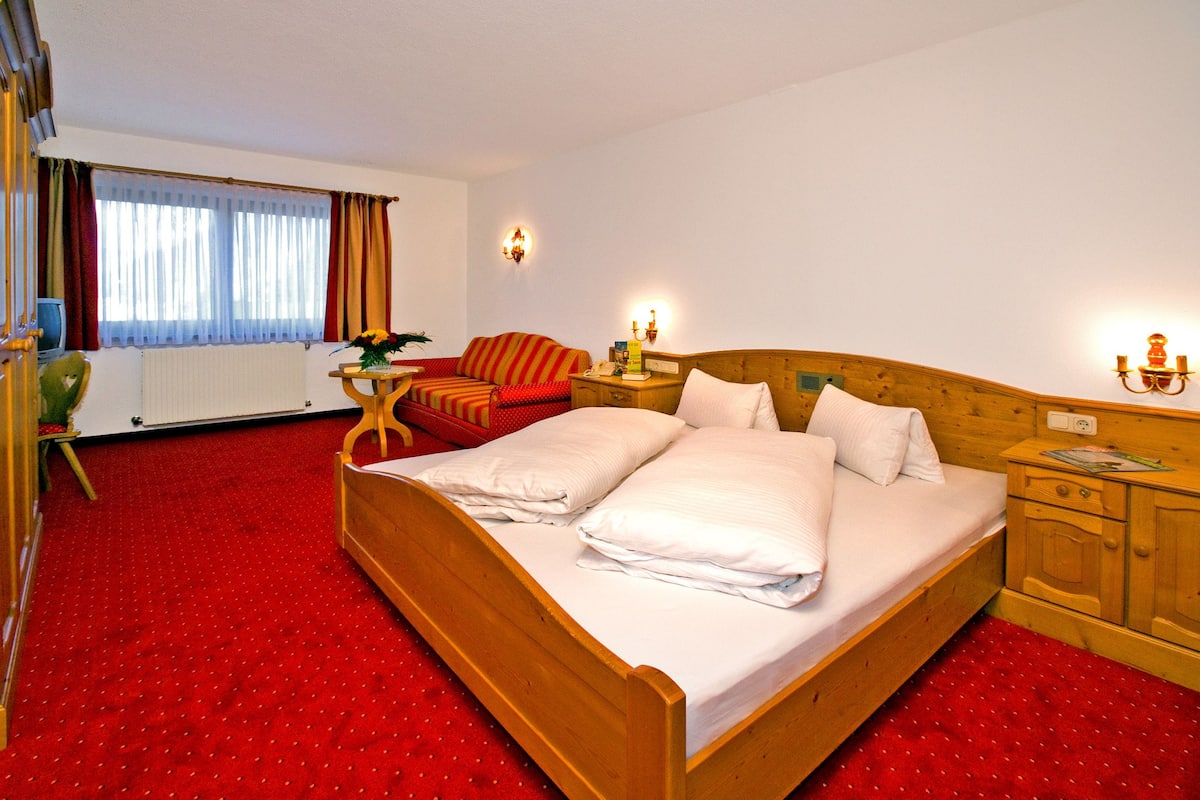 Ein Hotelzimmer mit einem großen Bett, einem Nachttisch aus Holz, einer Holzkommode, einem kleinen Tisch mit Stuhl und einem Fenster mit Vorhängen.