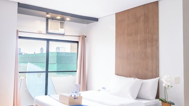 Una habitación de hotel moderna con una cama de madera, una mesita de noche negra, una planta en maceta y un ventanal con vistas a la ciudad.