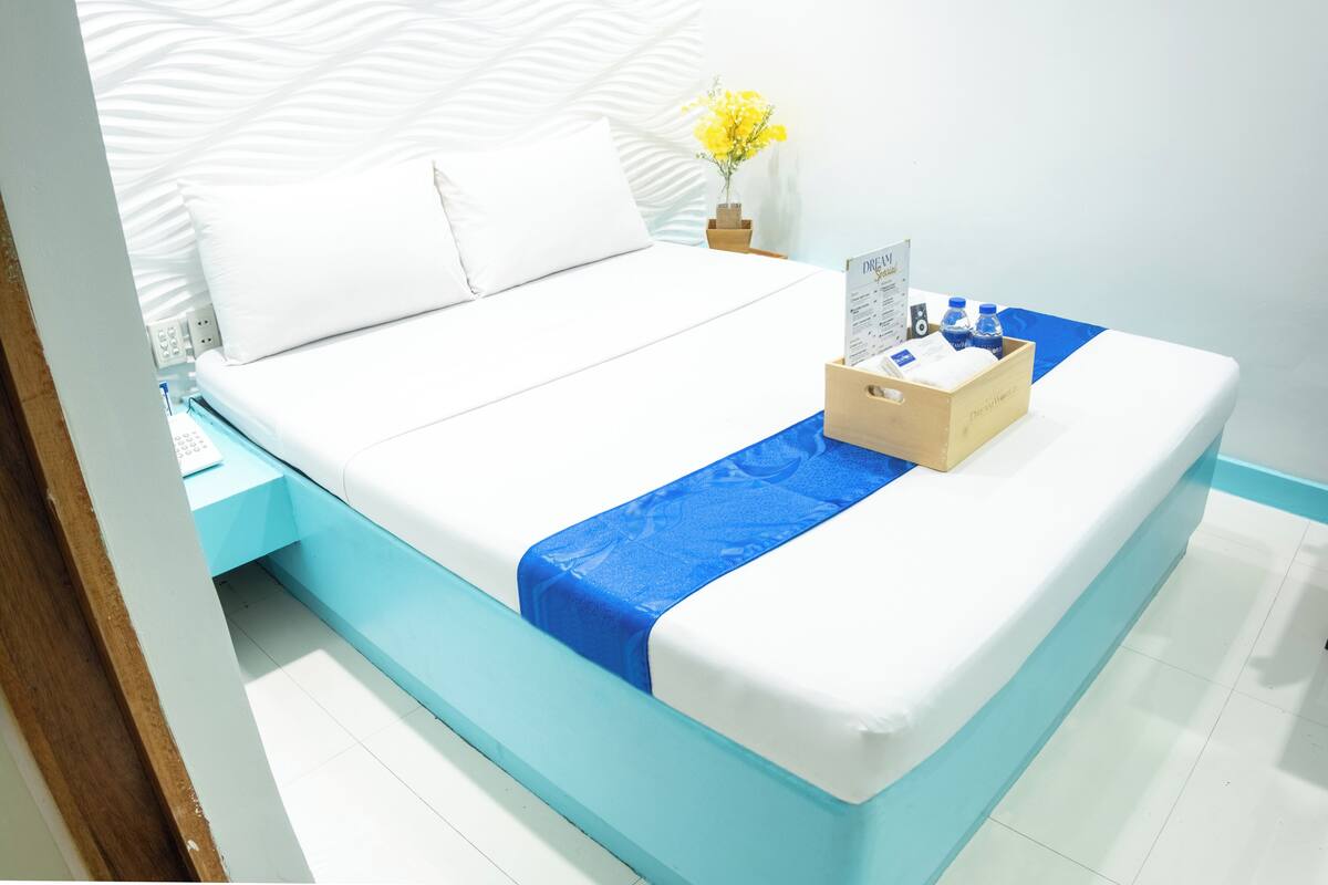Una cama bien hecha con un corredor azul, una mesita de noche de madera con un jarrón de flores amarillas y una pequeña mesa con un menú y agua embotellada.