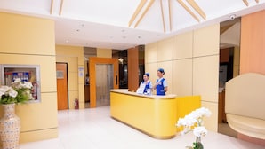 Zona de recepción de un hotel con un mostrador amarillo, dos recepcionistas, un jarrón grande con flores blancas y una planta en maceta sobre una mesa.