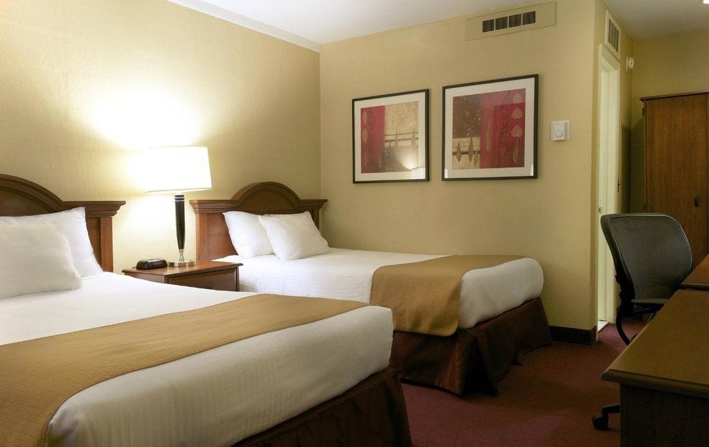 A hotel room with two beds, a desk, a chair, and two framed pictures on the wall.