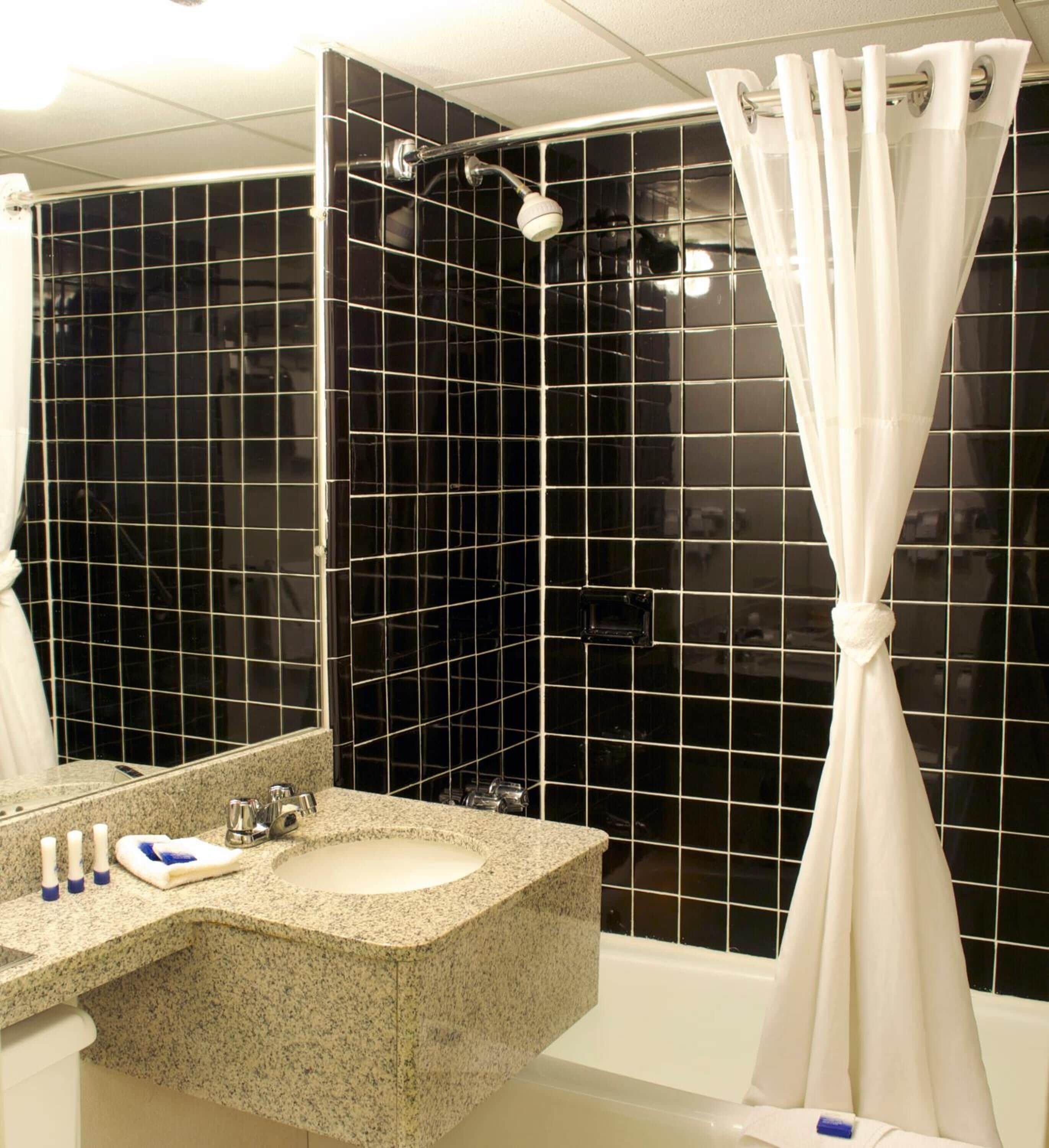 Une salle de bain avec un lavabo en granit, un miroir, une douche avec un rideau blanc et des murs recouverts de carreaux noirs.