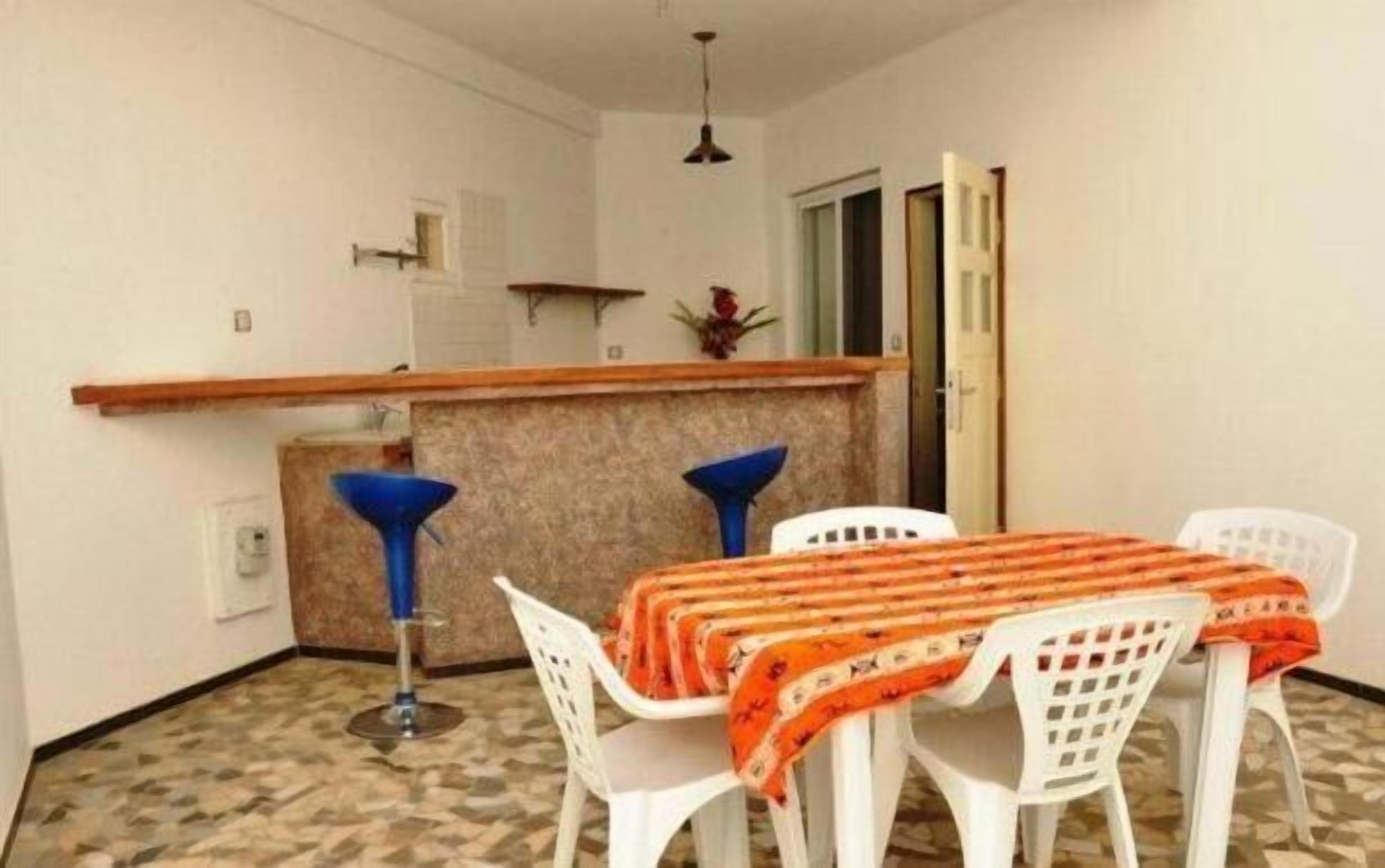 A small dining area with a table covered by a patterned tablecloth, two blue bar stools, and a white chair.